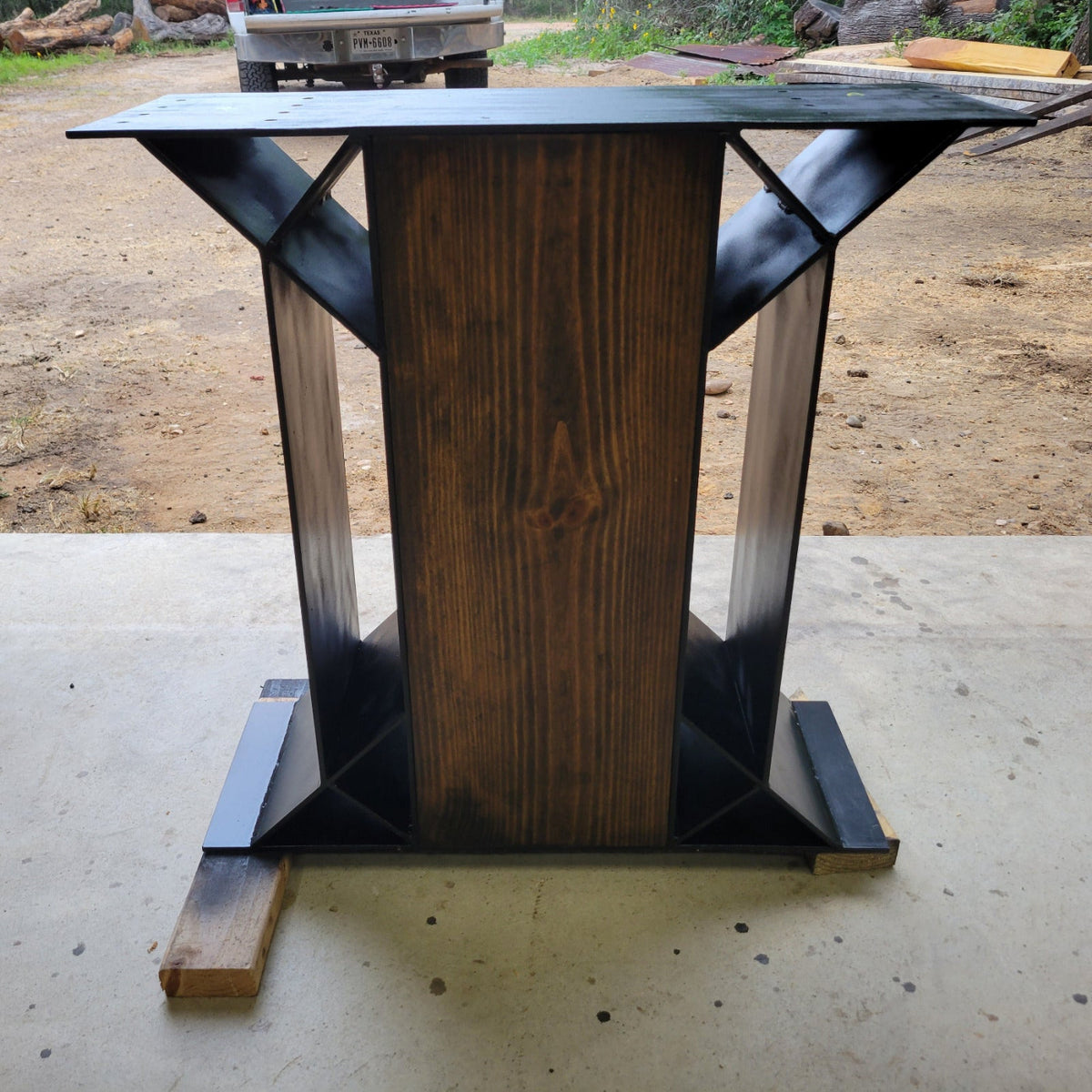 A rustic modern accent table with a dark wood central column and black metal geometric legs, shown on a concrete floor in an outdoor workshop setting.