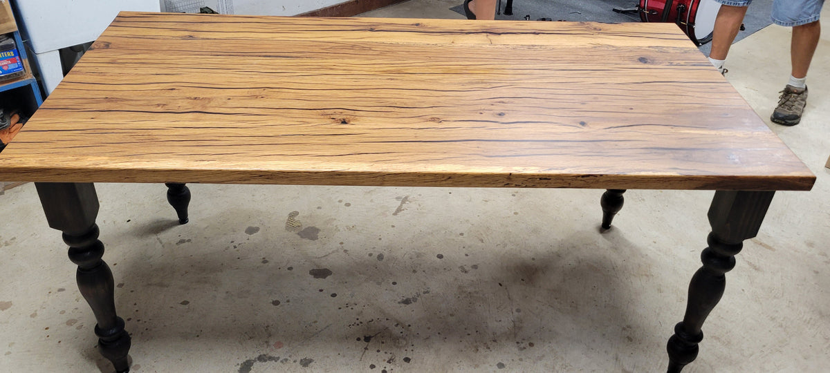 A rustic wooden dining table with a light brown wood top and dark turned legs, shown in a workshop setting.