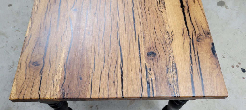A close-up view of a rustic wooden table top made from multiple natural wood planks with prominent dark grain lines, shown from above at a slight angle. The table is supported by dark legs on a concrete floor.