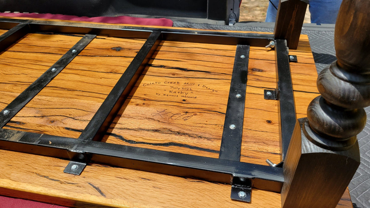 Close-up of a wooden table frame with black metal brackets and a turned wood spindle post, photographed outdoors on a paved surface.