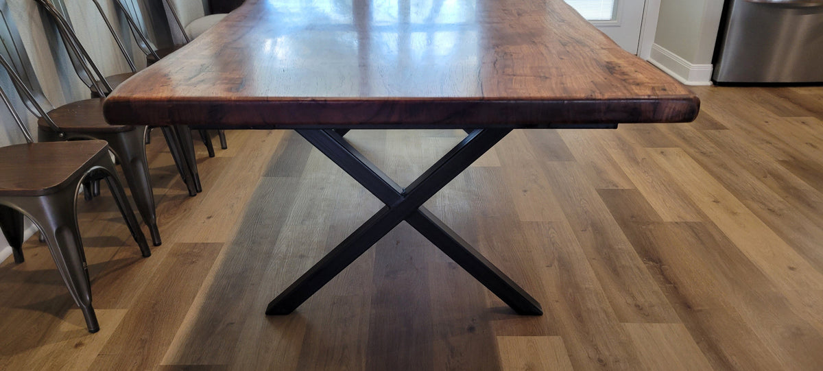 A large live-edge wooden dining table with a rich brown wood grain and natural bark edges, shown in a home or showroom setting with light wood floors.