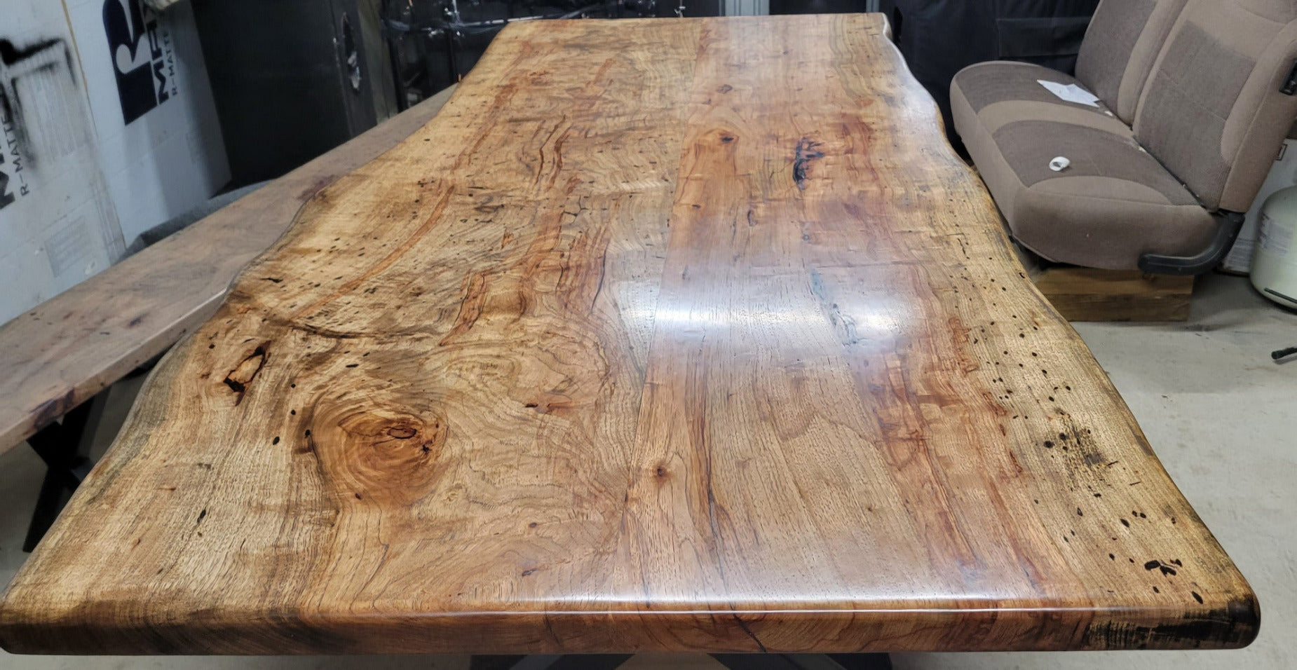 A rustic live-edge Pecan dining table with a natural wood top and black metal X-style legs, photographed in a workshop or warehouse setting.