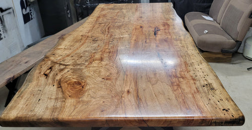 A rustic live-edge Pecan dining table with a natural wood top and black metal X-style legs, photographed in a workshop or warehouse setting.