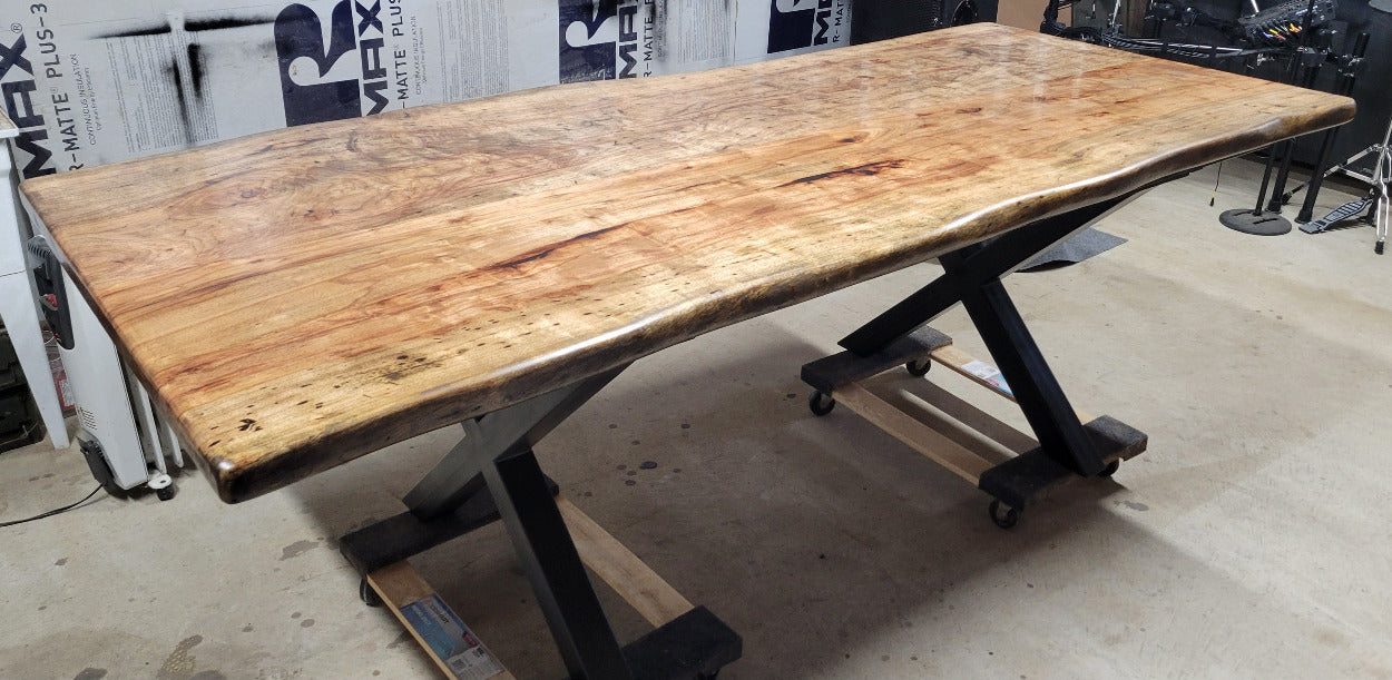 A rustic live-edge Pecan dining table with a natural wood top and black metal X-style legs, photographed in a workshop or warehouse setting.