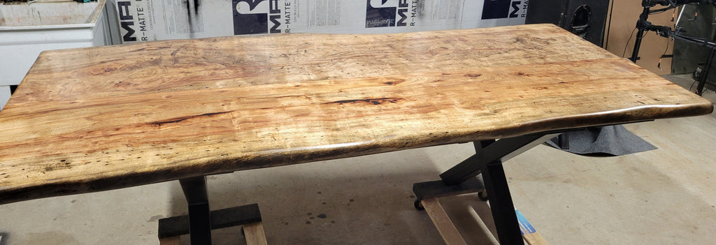 A rustic live-edge Pecan dining table with a natural wood top and black metal X-style legs, photographed in a workshop or warehouse setting.