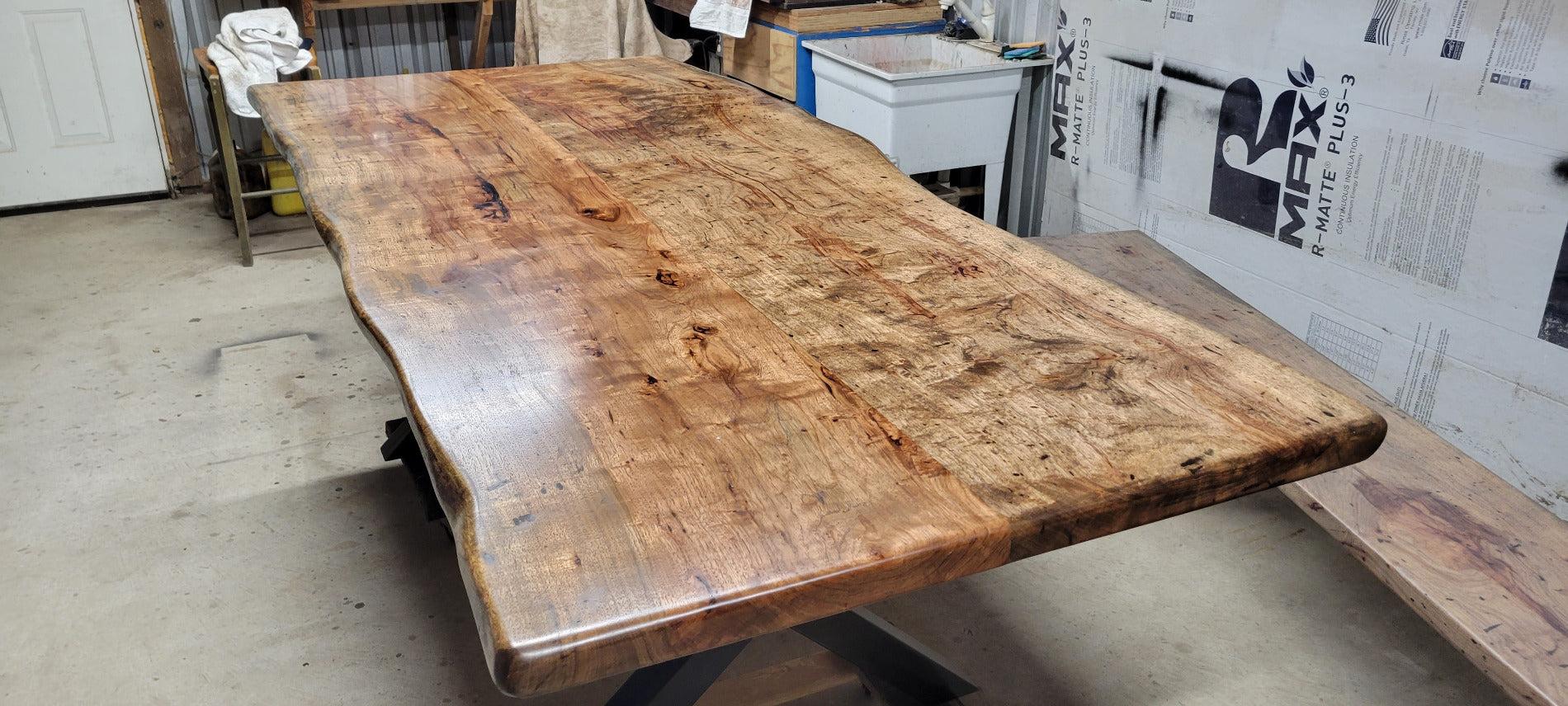 A rustic live-edge Pecan dining table with a natural wood top and black metal X-style legs, photographed in a workshop or warehouse setting.