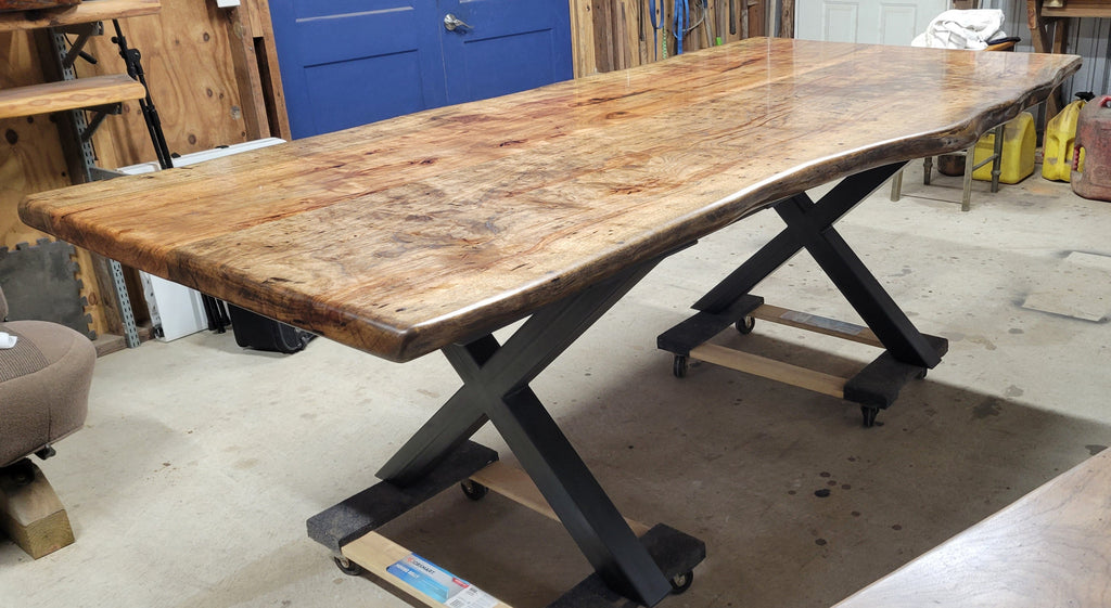 A rustic live-edge Pecan dining table with a natural wood top and black metal X-style legs, photographed in a workshop or warehouse setting.