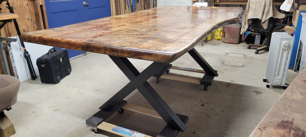 A rustic live-edge Pecan dining table with a natural wood top and black metal X-style legs, photographed in a workshop or warehouse setting.