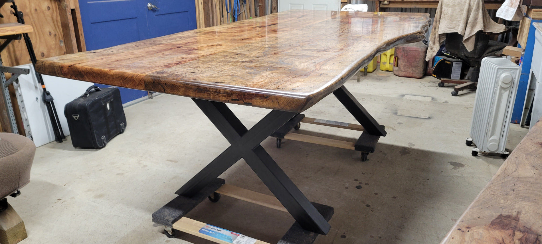 A rustic live-edge Pecan dining table with a natural wood top and black metal X-style legs, photographed in a workshop or warehouse setting.