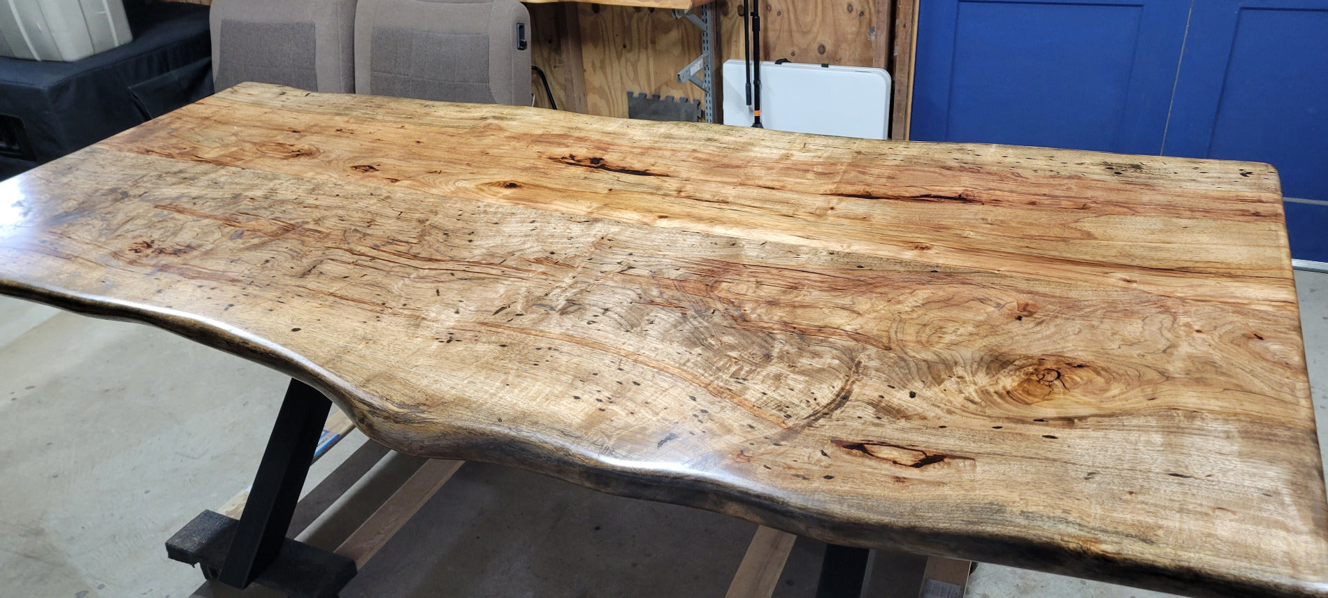 A rustic live-edge Pecan dining table with a natural wood top and black metal X-style legs, photographed in a workshop or warehouse setting.