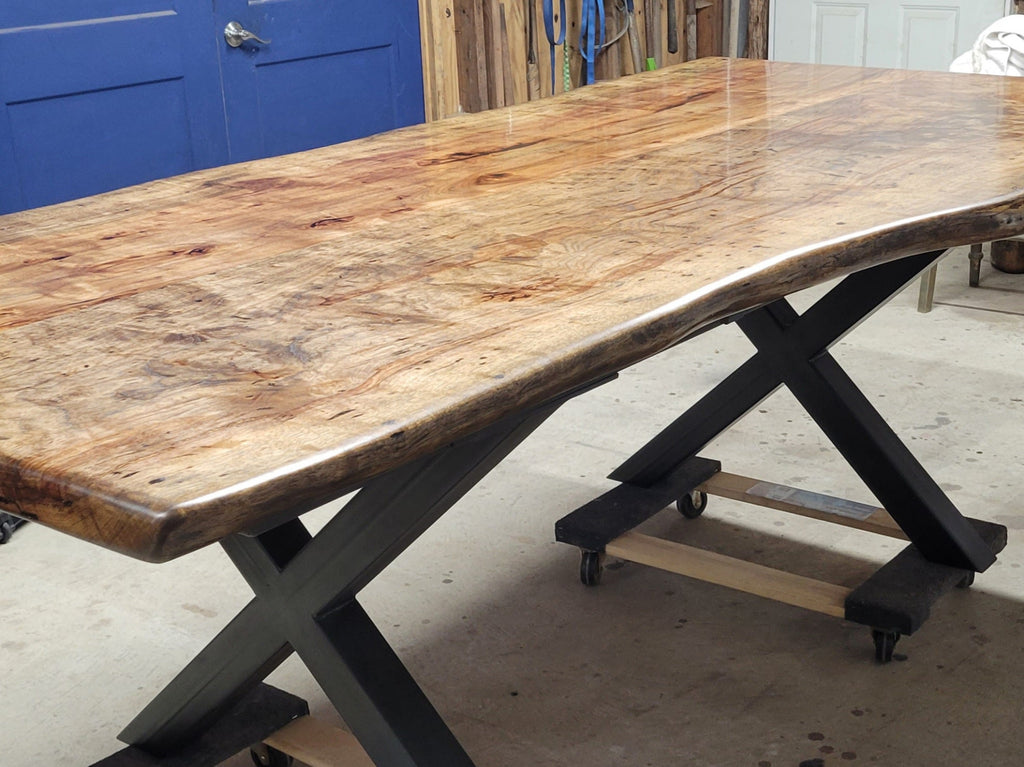 A rustic live-edge Pecan dining table with a natural wood top and black metal X-style legs, photographed in a workshop or warehouse setting.