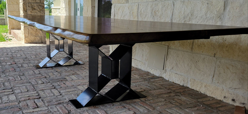 A long rectangular Pecan dining table with a dark wood top and black geometric metal trestle legs, shown on a brick patio beside a stone house exterior.