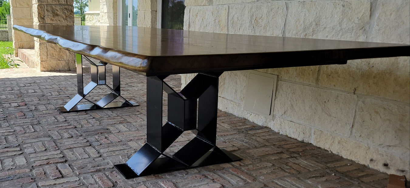 A long rectangular Pecan dining table with a dark wood top and black geometric metal trestle legs, shown on a brick patio beside a stone house exterior.
