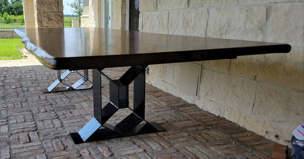 A long rectangular Pecan dining table with a dark wood top and black geometric metal trestle legs, shown on a brick patio beside a stone house exterior.