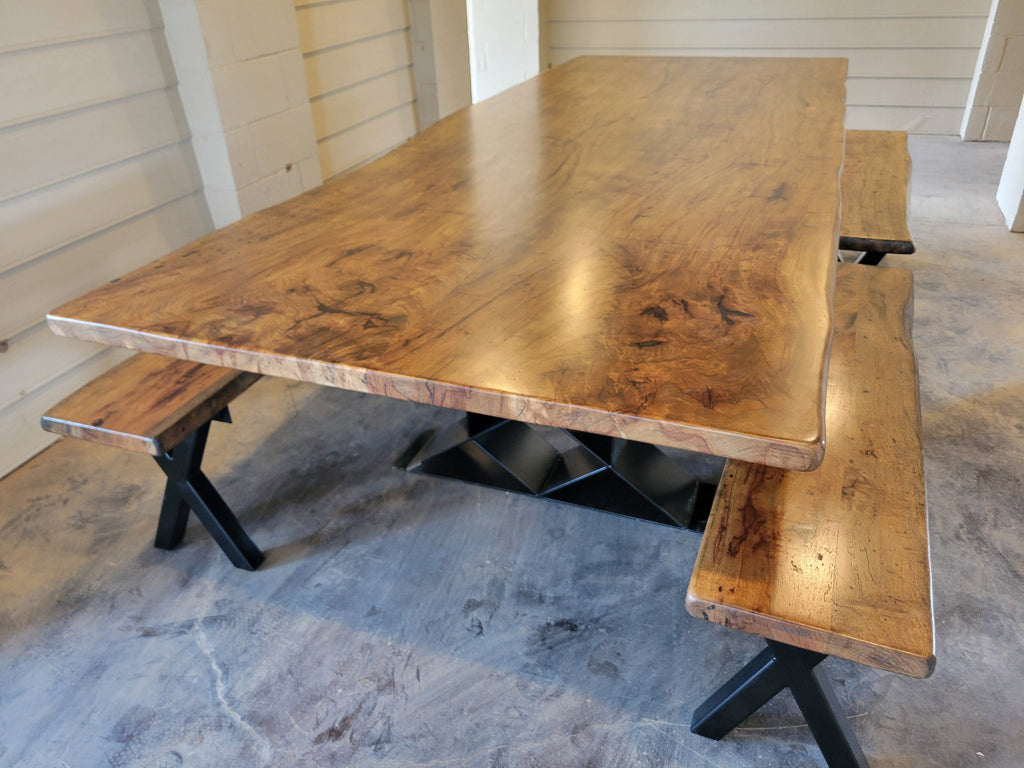 A rustic Pecan wooden dining table with a live edge and matching wooden benches on black metal A-frame legs, photographed with a concrete floor and light wood-paneled walls.
