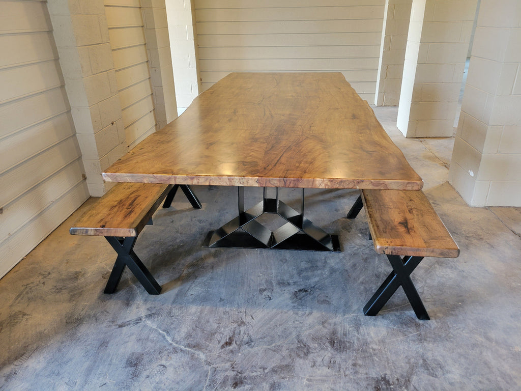 A rustic Pecan wooden dining table with a live edge and matching wooden benches on black metal A-frame legs, photographed with a concrete floor and light wood-paneled walls.