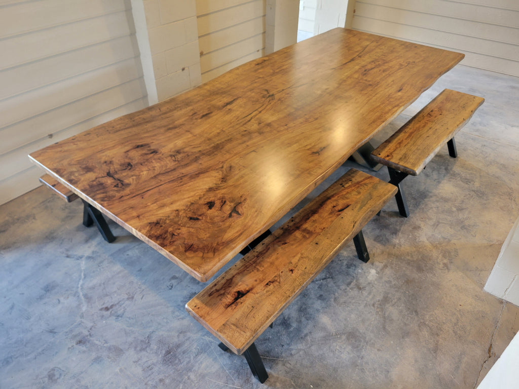A rustic Pecan wooden dining table with a live edge and matching wooden benches on black metal A-frame legs, photographed with a concrete floor and light wood-paneled walls.