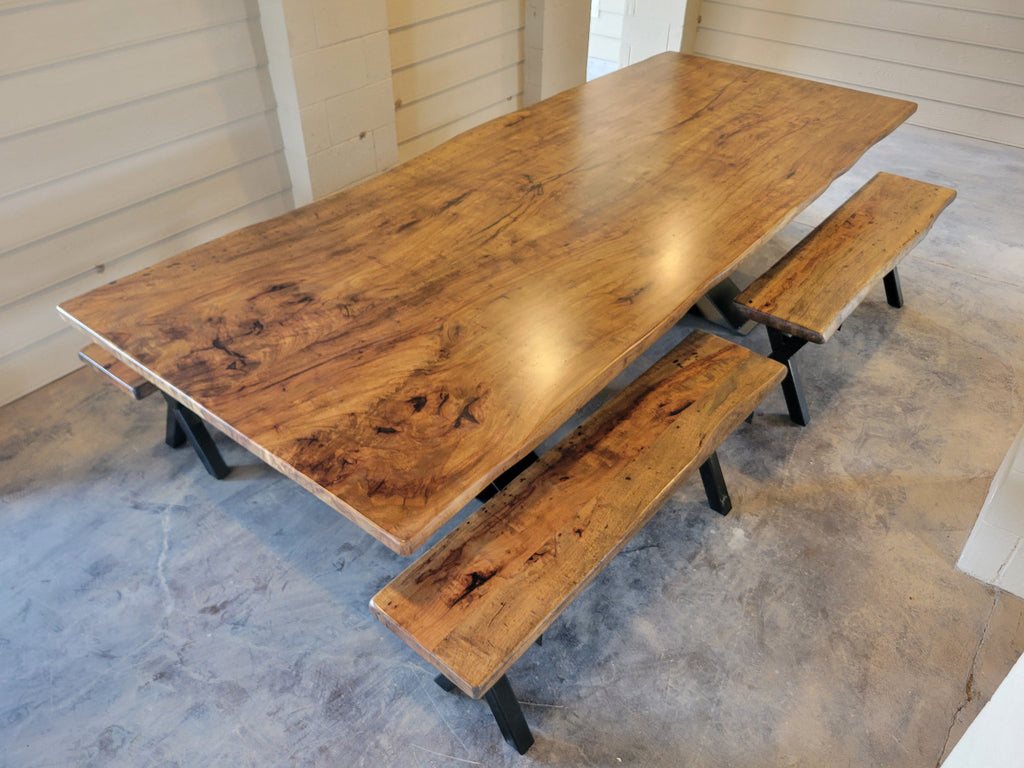 A rustic Pecan wooden dining table with a live edge and matching wooden benches on black metal A-frame legs, photographed with a concrete floor and light wood-paneled walls.