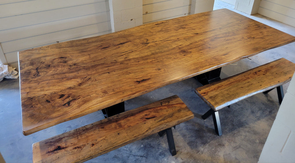 A rustic Pecan wooden dining table with a live edge and matching wooden benches on black metal A-frame legs, photographed with a concrete floor and light wood-paneled walls.