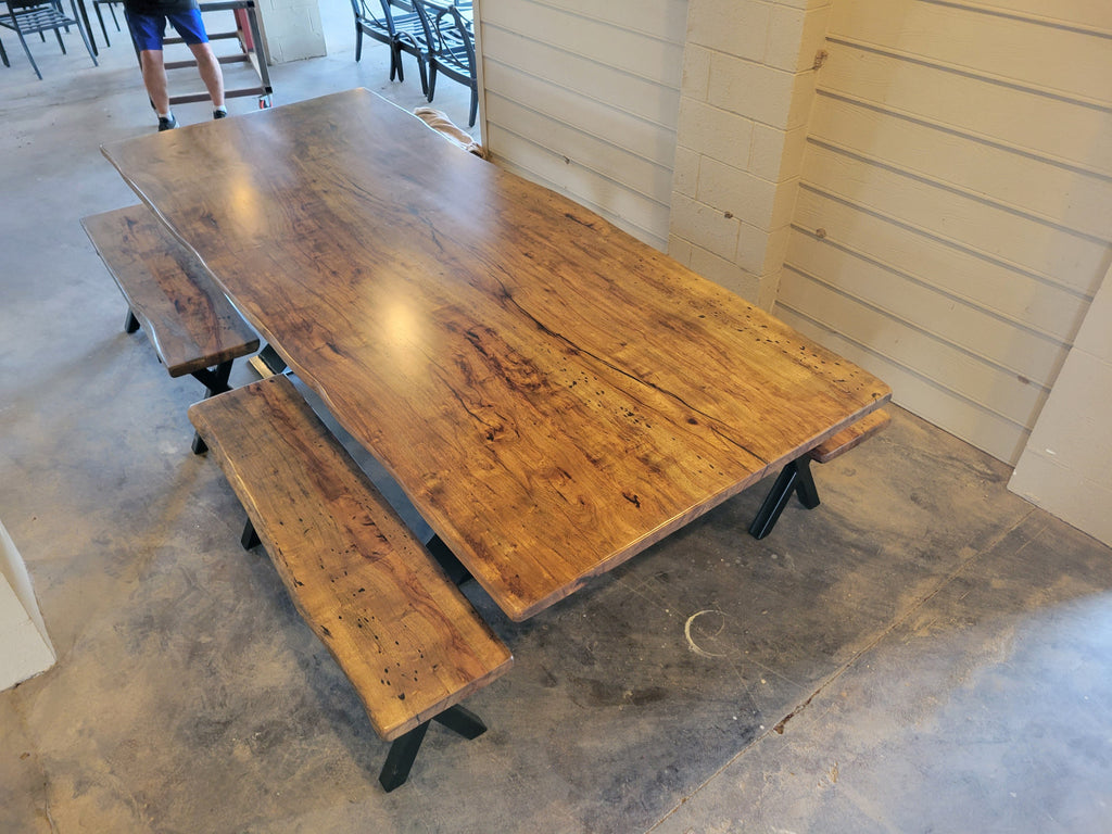 A rustic Pecan wooden dining table with a live edge and matching wooden benches on black metal A-frame legs, photographed with a concrete floor and light wood-paneled walls.