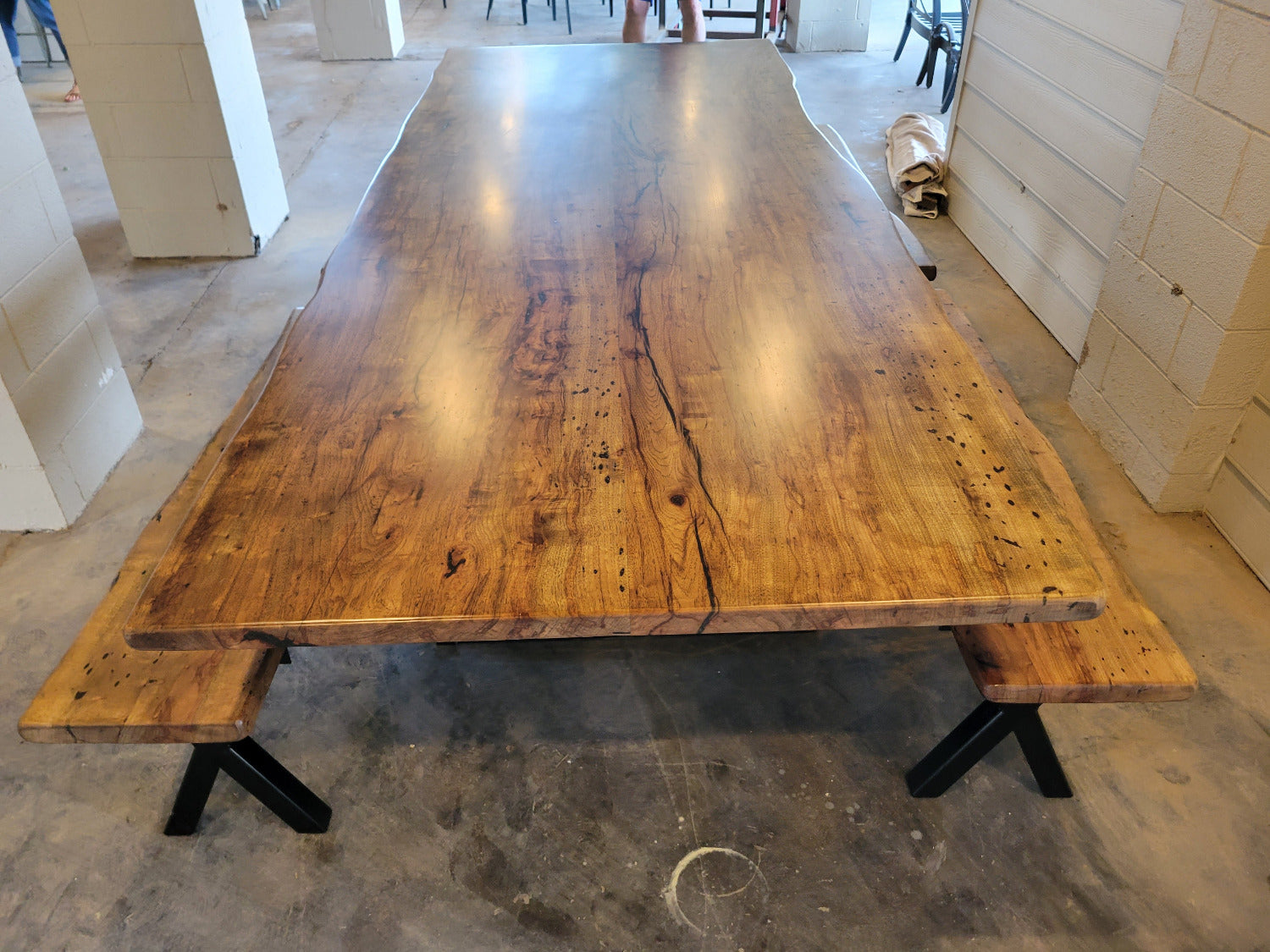 A rustic Pecan wooden dining table with a live edge and matching wooden benches on black metal A-frame legs, photographed with a concrete floor and light wood-paneled walls.