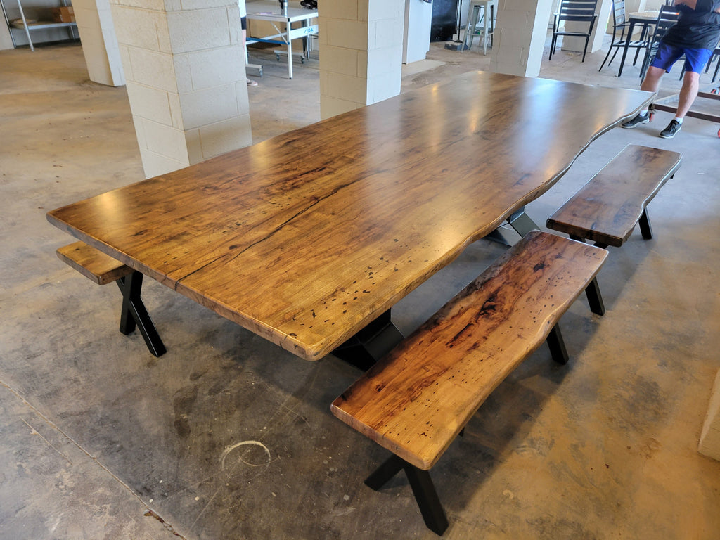 A rustic Pecan wooden dining table with a live edge and matching wooden benches on black metal A-frame legs, photographed with a concrete floor and light wood-paneled walls.