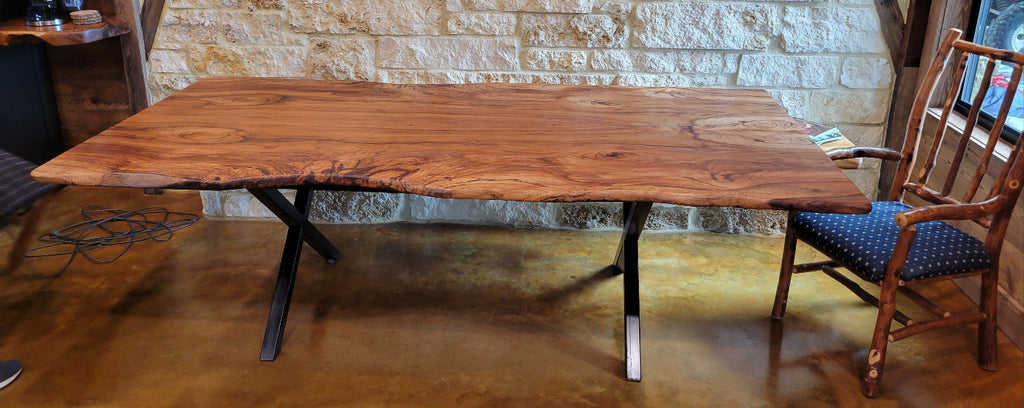 A rustic Pecan live-edge wooden dining table with black metal legs, shown with a stone wall background and wooden chairs.