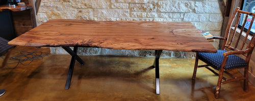 A rustic Pecan live-edge wooden dining table with black metal legs, shown with a stone wall background and wooden chairs.