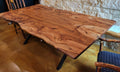 A rustic Pecan live-edge wooden dining table with black metal legs, shown in a restaurant or cafe setting with a stone wall background and wooden chairs.