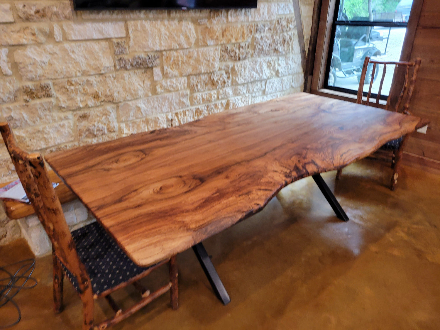 A rustic Pecan live-edge wooden dining table with black metal legs, shown with a stone wall background and wooden chairs.
