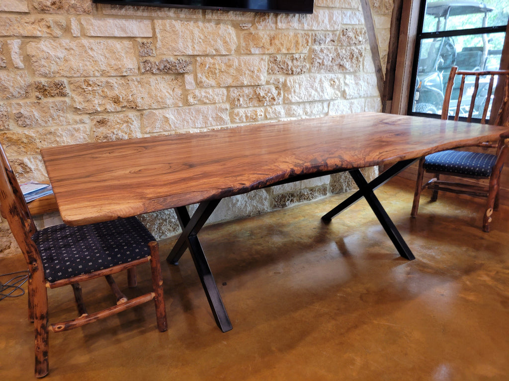 A rustic Pecan live-edge wooden dining table with black metal legs, shown with a stone wall background and wooden chairs.