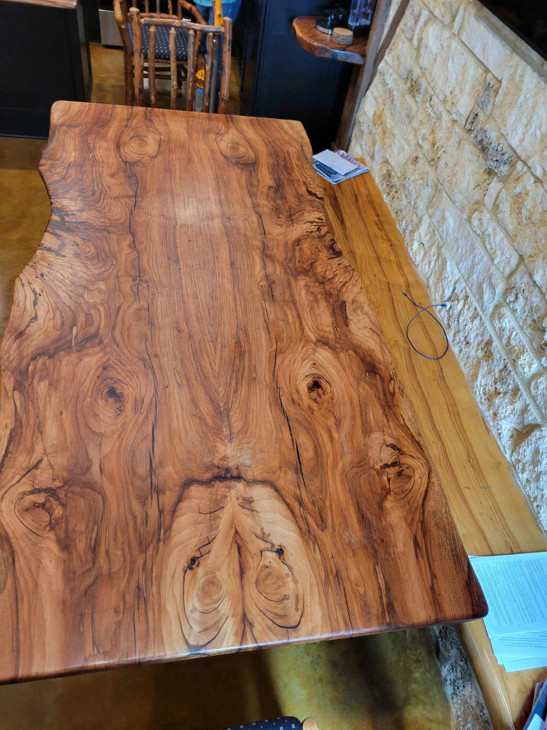A rustic Pecan live-edge wooden dining table with black metal legs, shown with a stone wall background and wooden chairs.