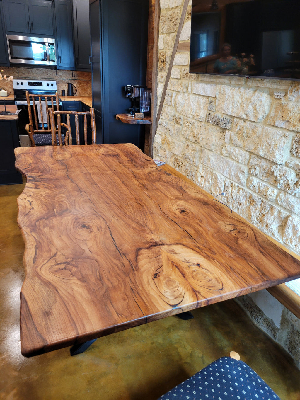 A rustic Pecan live-edge wooden dining table with black metal legs, shown with a stone wall background and wooden chairs.