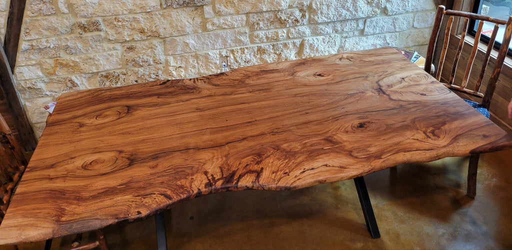 A rustic Pecan live-edge wooden dining table with black metal legs, shown with a stone wall background and wooden chairs.