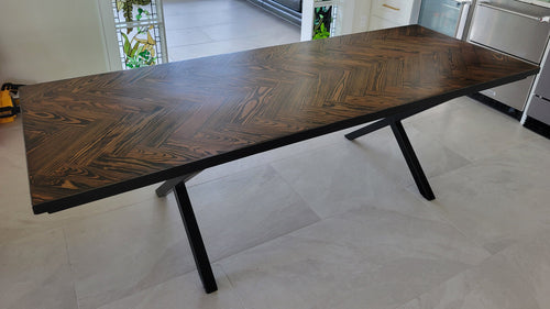 A rectangular long leaf pine dining table with a dark brown herringbone pattern top and black metal legs and metal sides shown in a home interior setting.