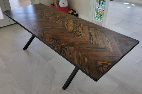 A rectangular long leaf pine dining table with a dark brown herringbone pattern top and black metal legs and metal sides shown in a home interior setting.