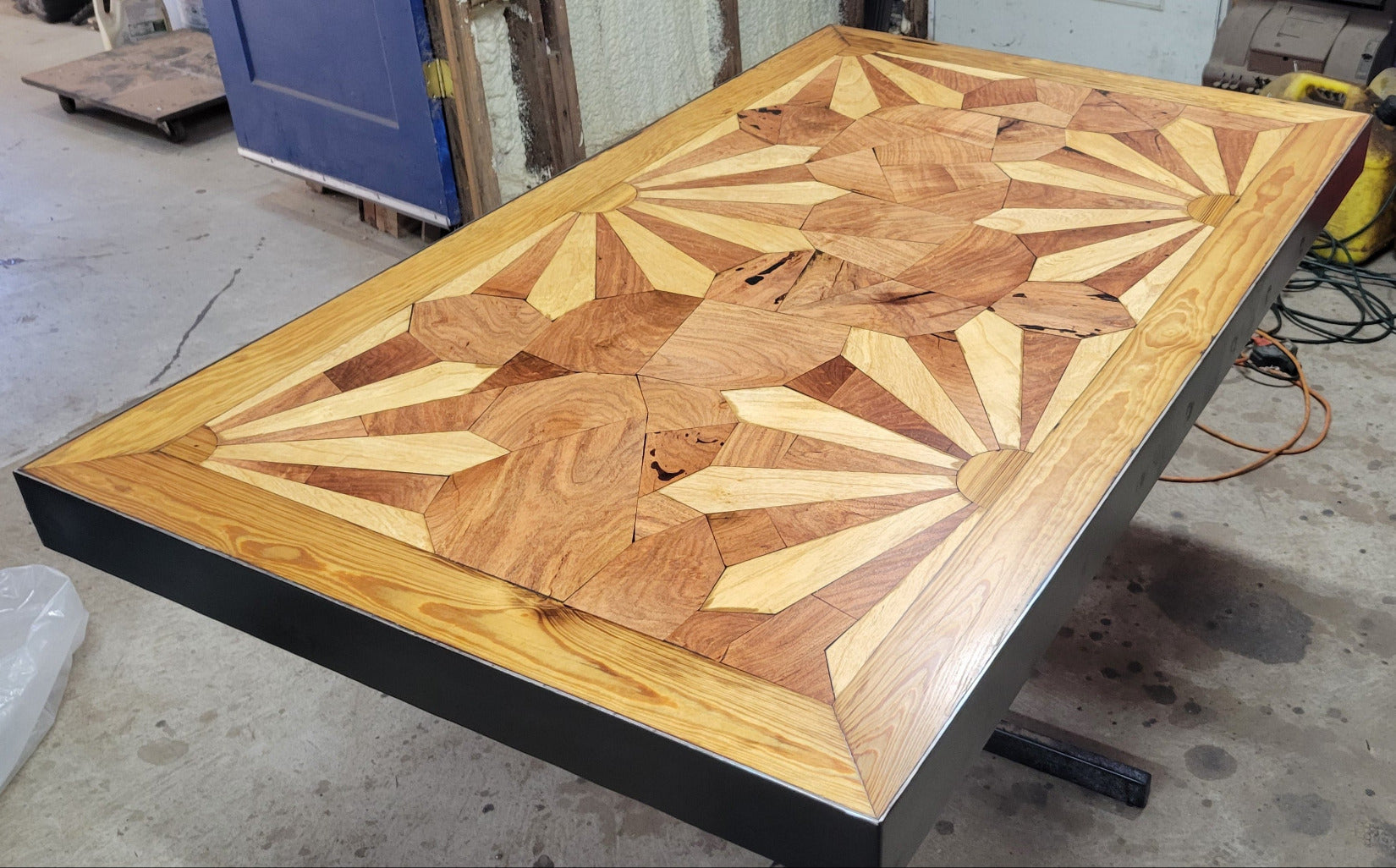 A rectangular wooden dining table with a black metal pedestal base, set in a wooden booth or cafe setting. The tabletop features a detailed geometric inlay pattern in shades of light and dark brown wood made from Ash and mesquite wood