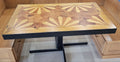A rectangular wooden dining table with a black metal pedestal base, set in a wooden booth or cafe setting. The tabletop features a detailed geometric inlay pattern in shades of light and dark brown wood made from Ash and mesquite wood