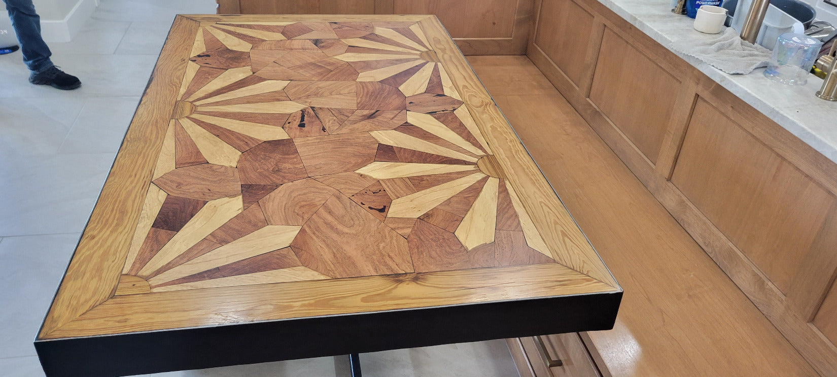 A rectangular wooden dining table with a black metal pedestal base, set in a wooden booth or cafe setting. The tabletop features a detailed geometric inlay pattern in shades of light and dark brown wood made from Ash and mesquite wood