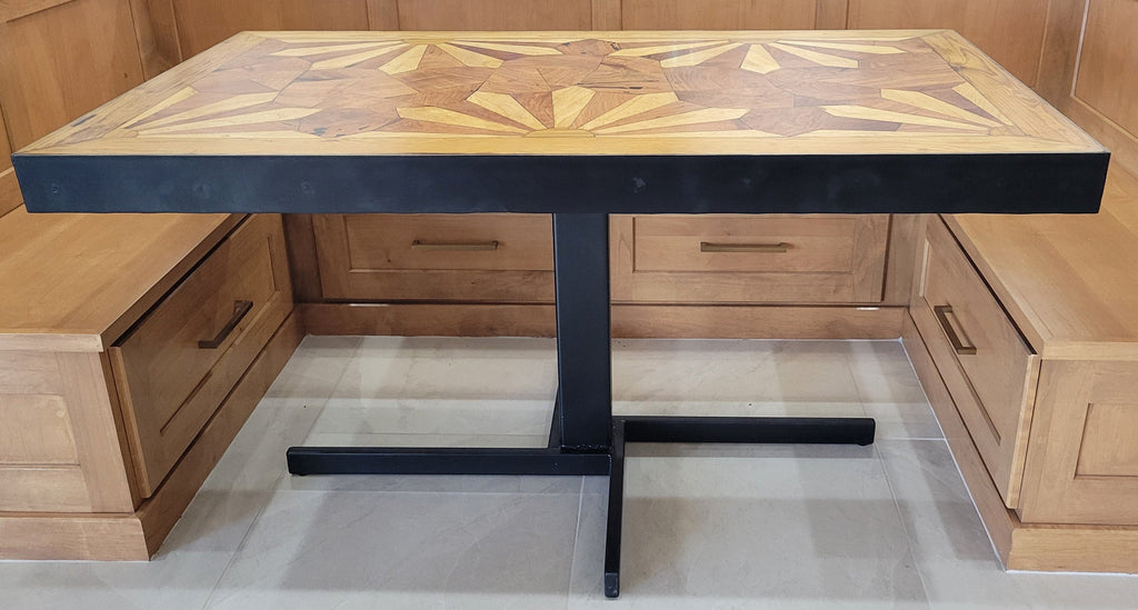 A rectangular wooden dining table with a black metal pedestal base, set in a wooden booth or cafe setting. The tabletop features a detailed geometric inlay pattern in shades of light and dark brown wood made from Ash and mesquite wood