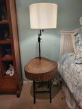A rustic wooden table made from a live-edge tree slab with built in lamp stands and white drum lamp, black metal custom stand and turquoise epoxy fill
