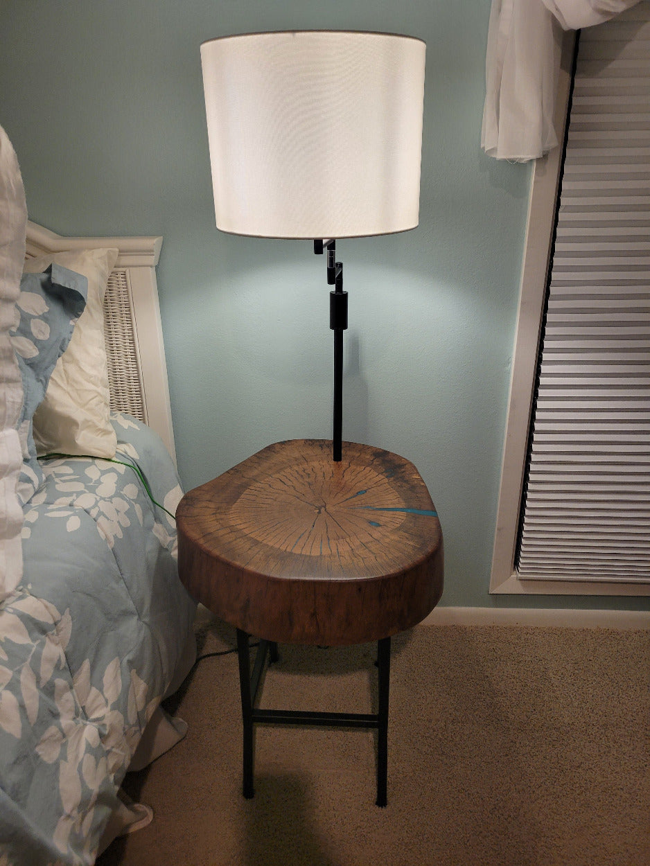 A rustic wooden table made from a live-edge tree slab with built in lamp stands and white drum lamp, black metal custom stand and turquoise epoxy fill