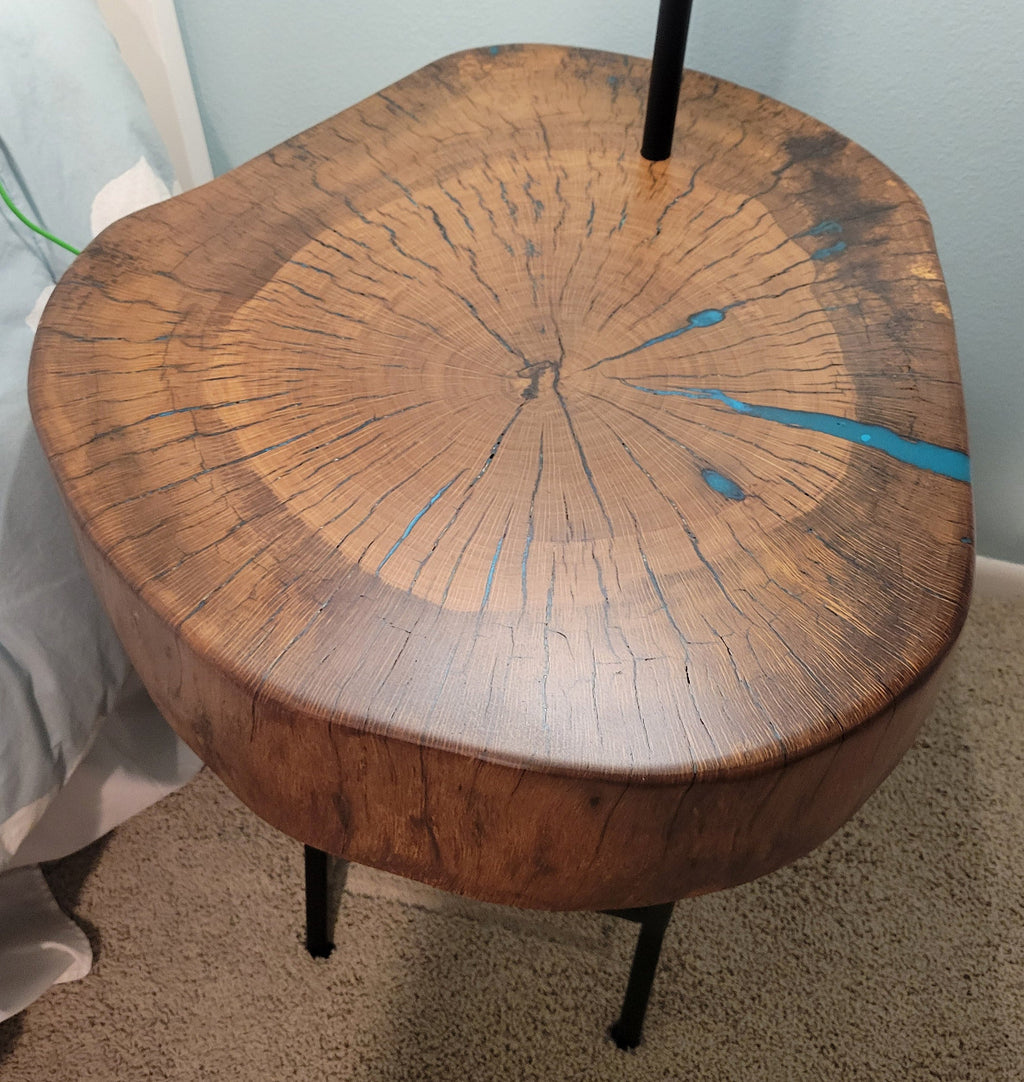 A rustic wooden table made from a live-edge tree slab with built in lamp stands and white drum lamp, black metal custom stand and turquoise epoxy fill