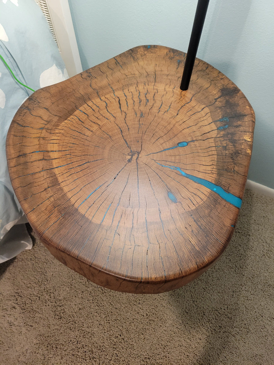 A rustic wooden table made from a live-edge tree slab with built in lamp stands and white drum lamp, black metal custom stand and turquoise epoxy fill