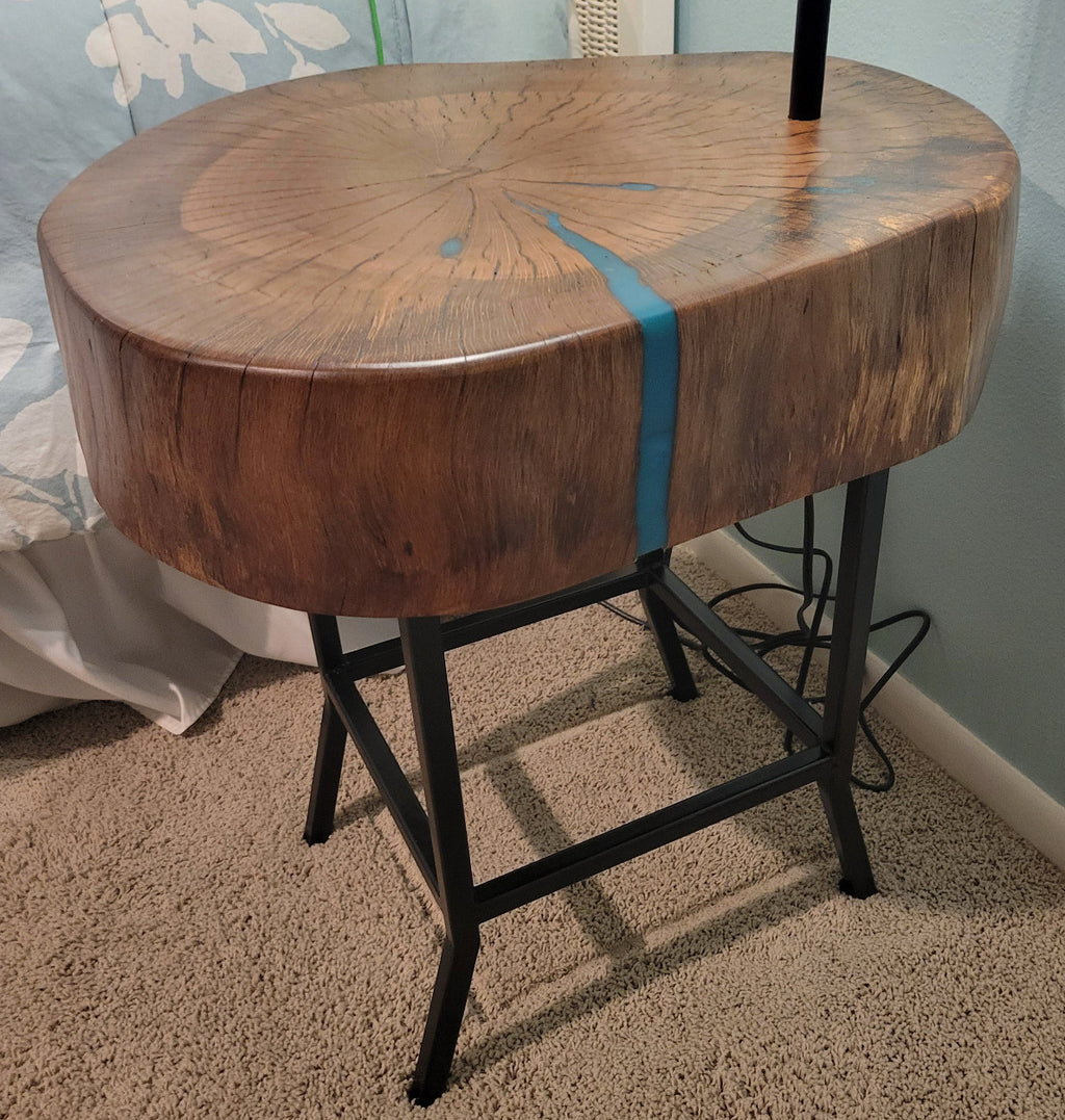 A rustic wooden table made from a live-edge tree slab with built in lamp stands and white drum lamp, black metal custom stand and turquoise epoxy fill