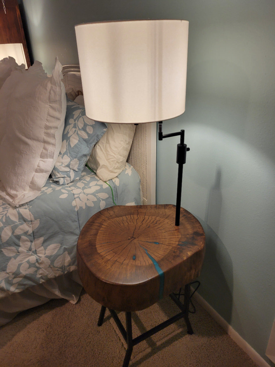 A rustic wooden table made from a live-edge tree slab with built in lamp stands and white drum lamp, black metal custom stand and turquoise epoxy fill