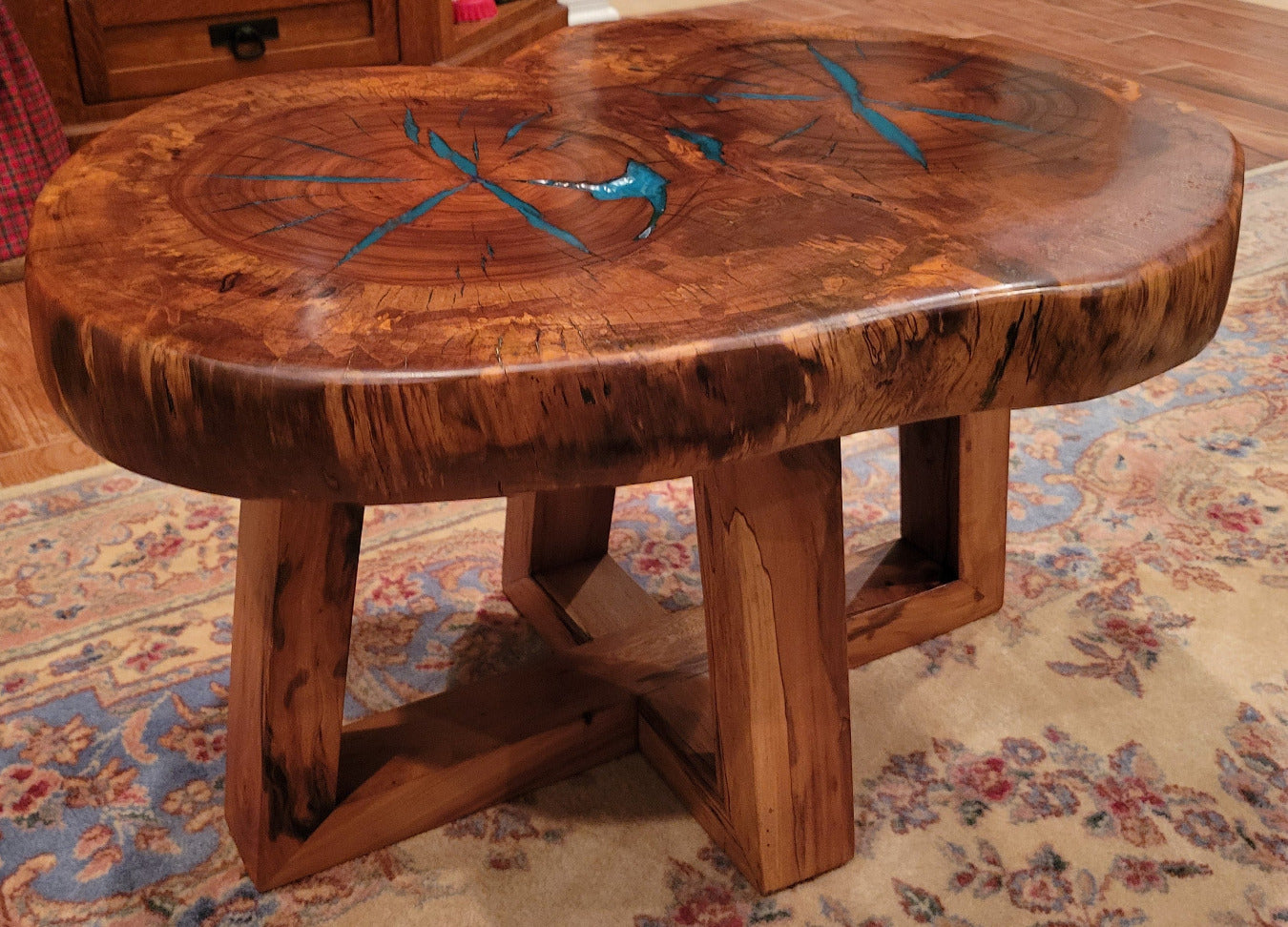 A rustic, organic-shaped wooden coffee table with a rich brown wood grain and bright turquoise blue resin inlay details, shown on a patterned oriental rug.