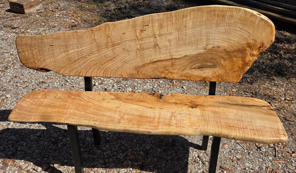 A rustic Ash live-edge wooden bench with a thick natural wood backrest and seat supported by black metal legs, photographed outdoors on gravel.