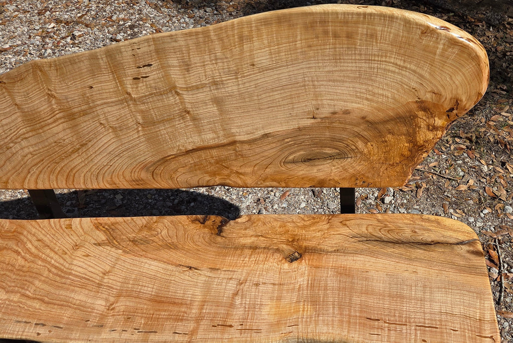 A rustic Ash live-edge wooden bench with a thick natural wood backrest and seat supported by black metal legs, photographed outdoors on gravel.