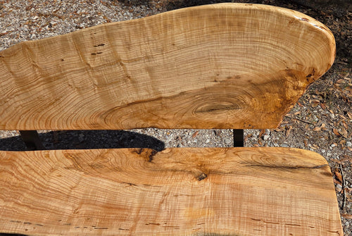 A rustic Ash live-edge wooden bench with a thick natural wood backrest and seat supported by black metal legs, photographed outdoors on gravel.